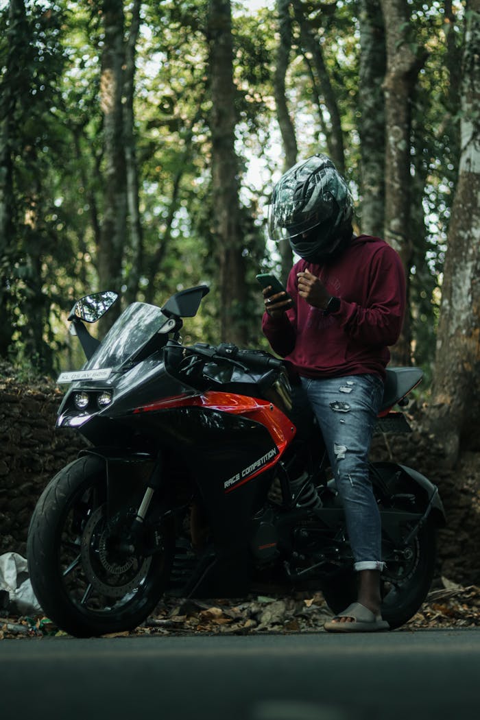 Man on motorbike wearing helmet, using phone amidst forest background.