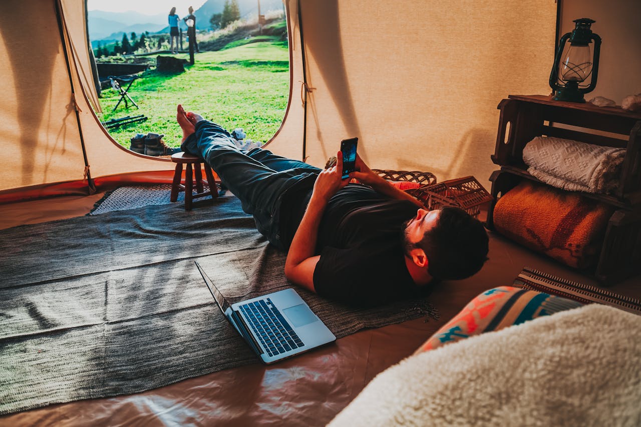 Man relaxing in a tent using smartphone and laptop, depicting a mix of nature and technology.