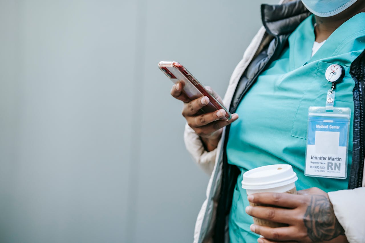 Nurse in scrubs checking phone while holding a takeaway coffee outdoors.