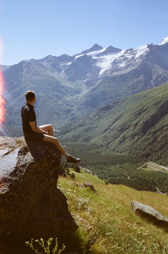 A person sits on a rock, admiring a stunning view of snow-capped mountains and a lush valley in summer.